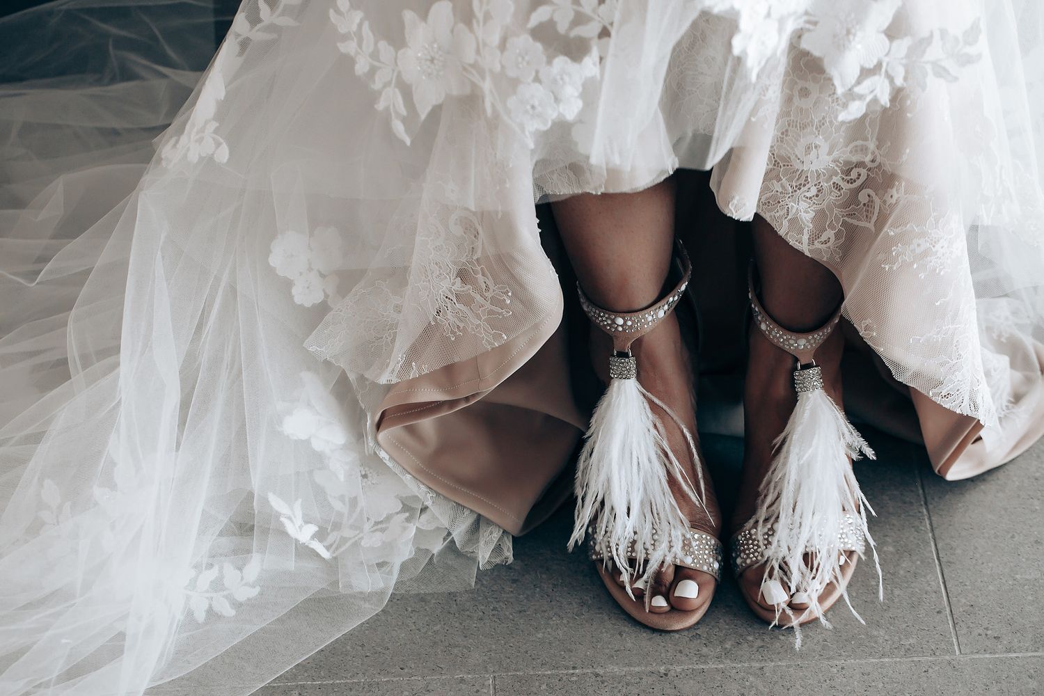 Close-up details of elegant bridal shoes with feather embellishments peeking out from under a wedding dress.