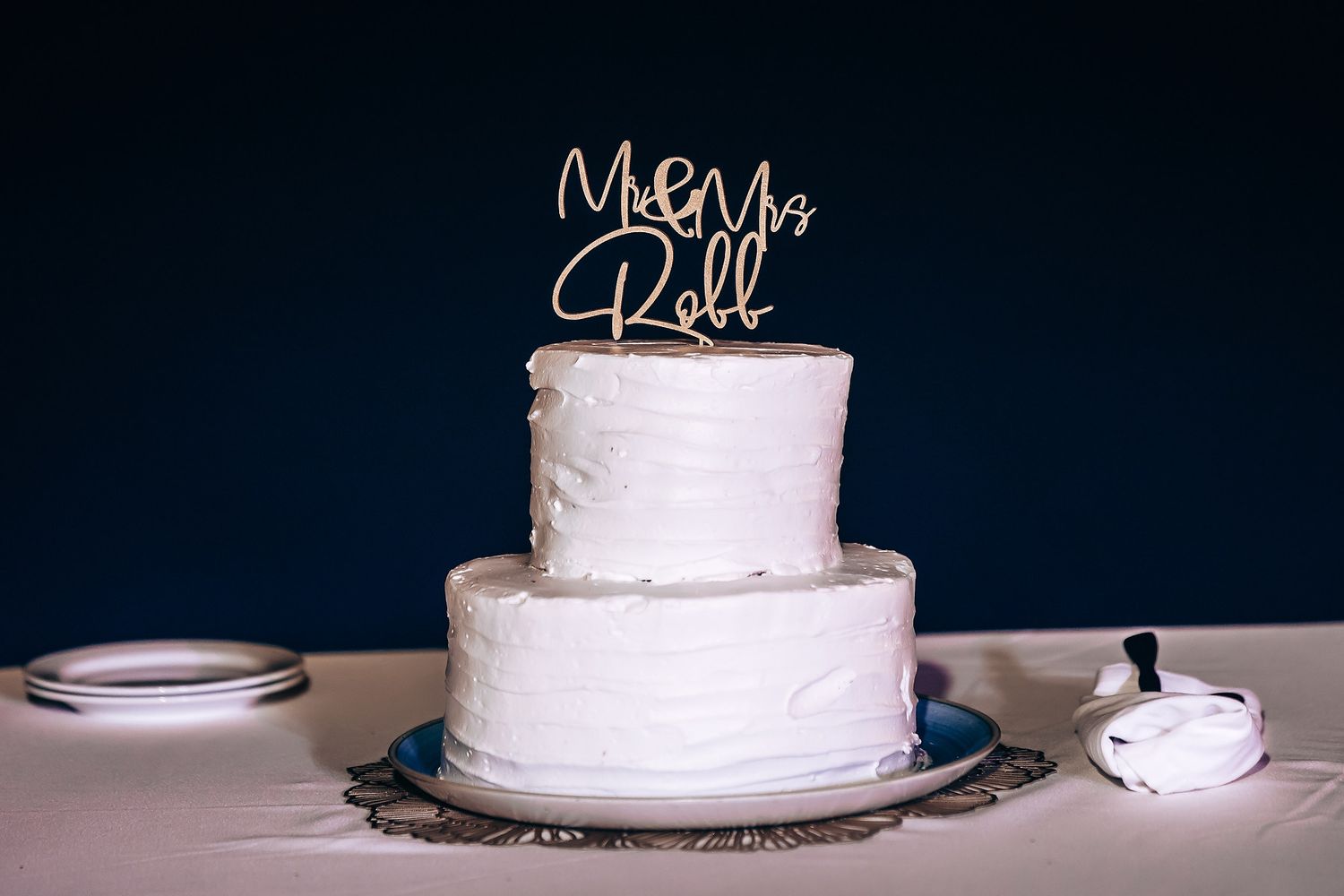 White tiered wedding cake with metallic name topper on dark background.