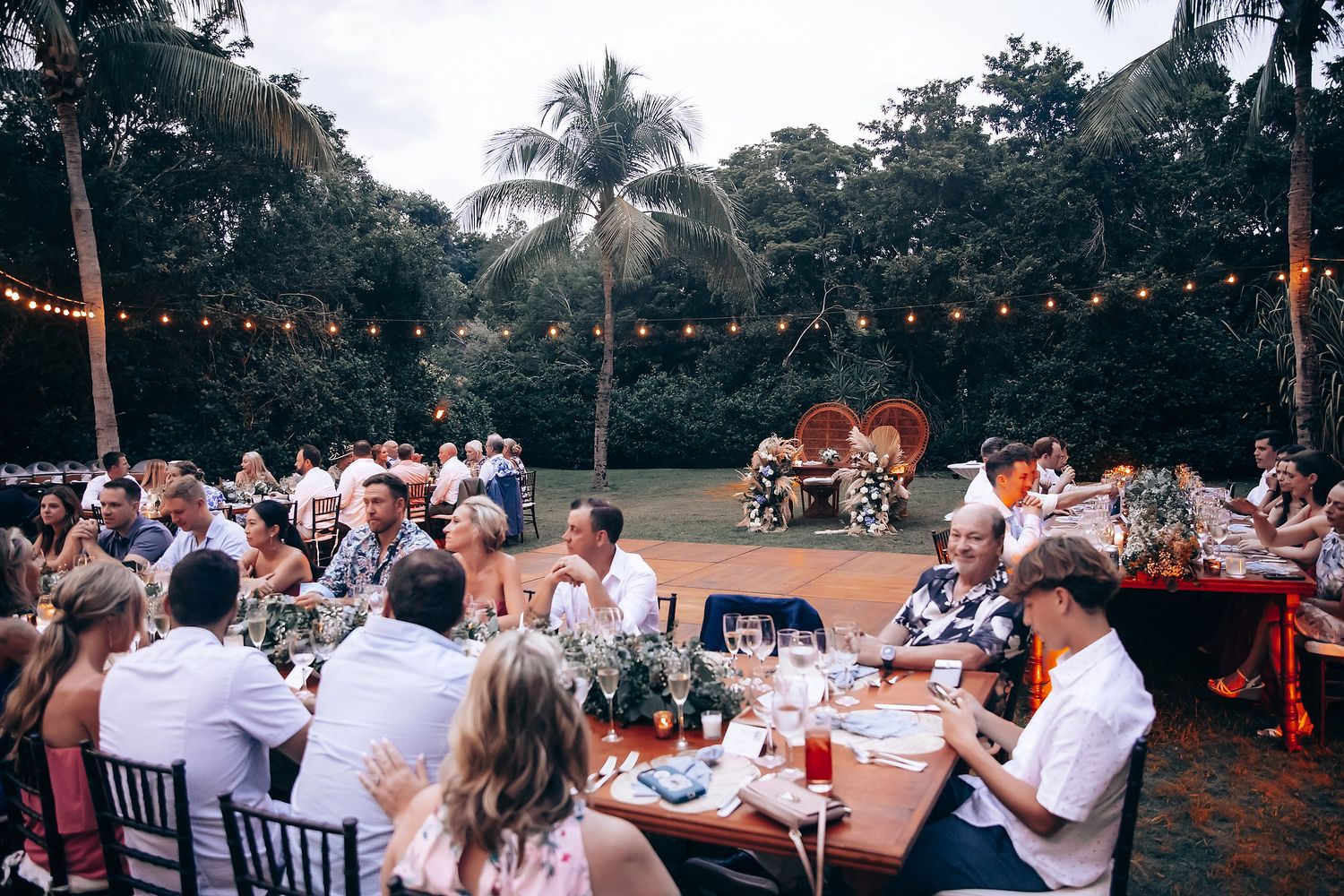 Wedding reception dinner under string lights with palm trees at sunset.