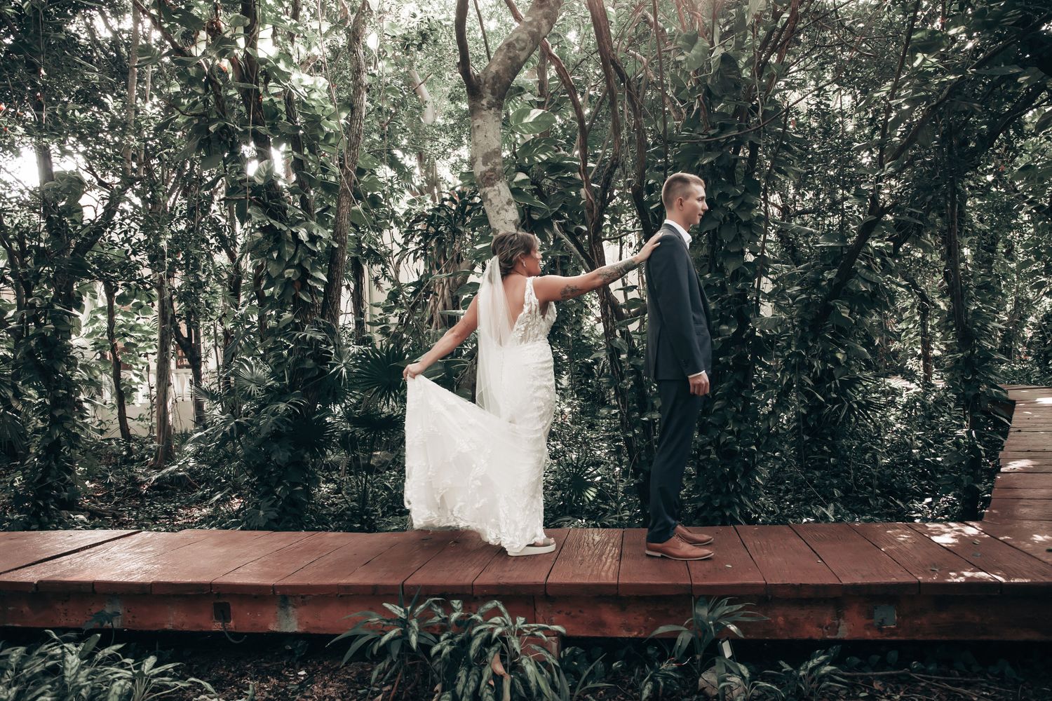 Bride and groom hold hands on a wooden deck surrounded by lush tropical forest during their wedding.