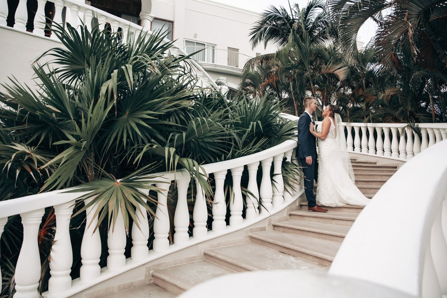 Bride and groom embracing on grand white staircase surrounded by tropical palm trees.