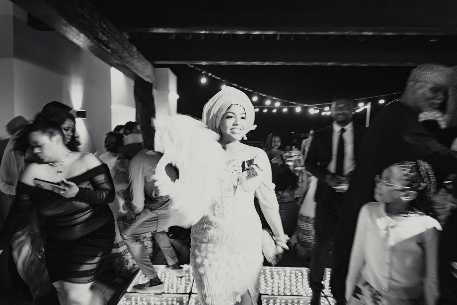 Black and white photo of bride dancing joyfully with guests under string lights at wedding reception.