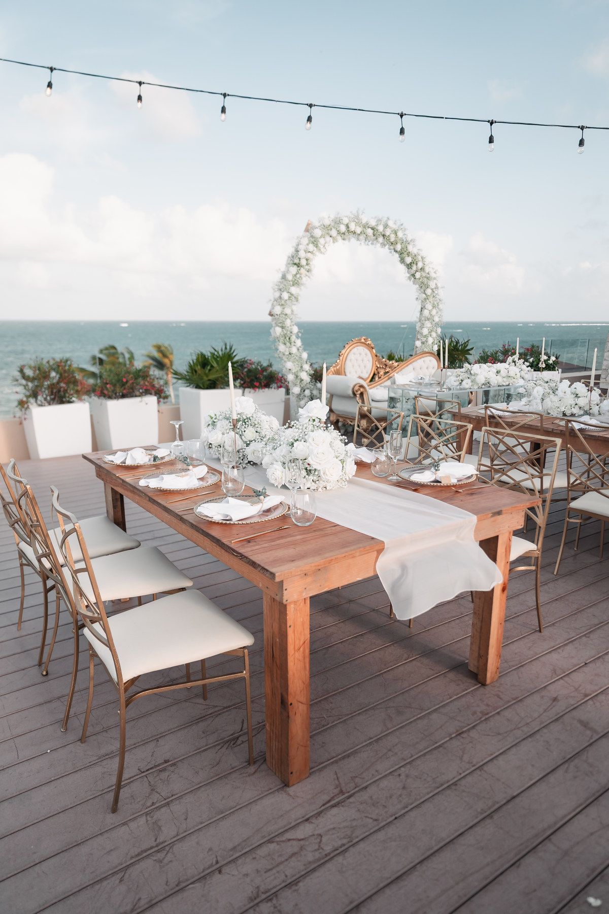 Elegant outdoor wedding reception table with white floral arch, string lights, and ocean views.