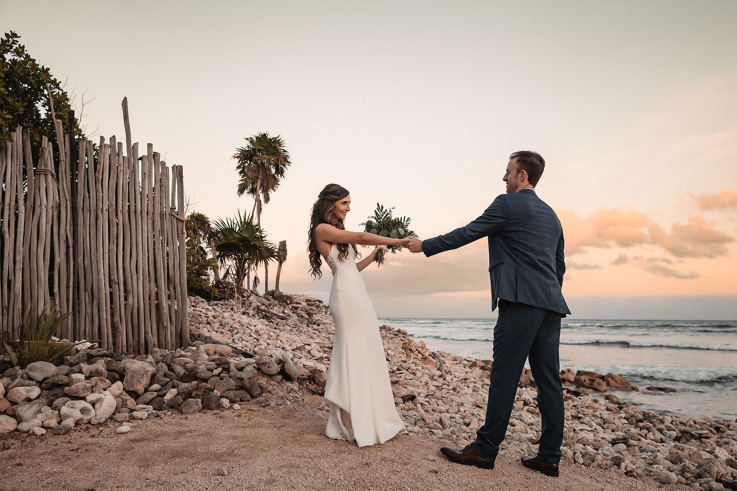 Bride and groom hold hands on rocky beach at sunset during wedding.