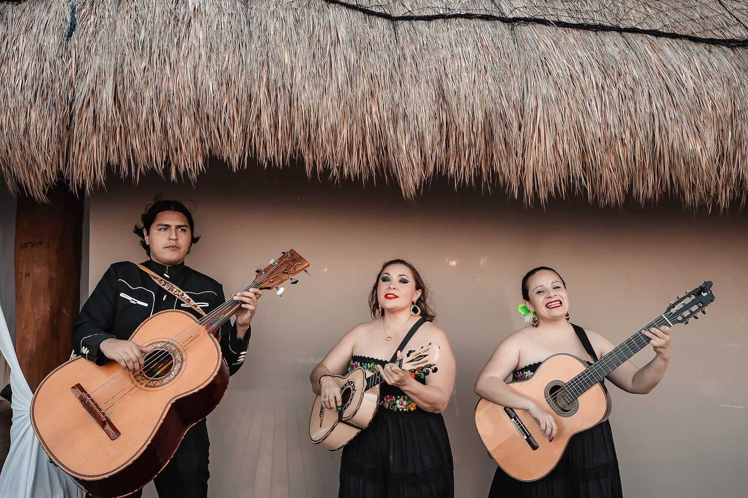 Three mariachi musicians in black outfits playing guitars under a thatched roof at an outdoor venue.
