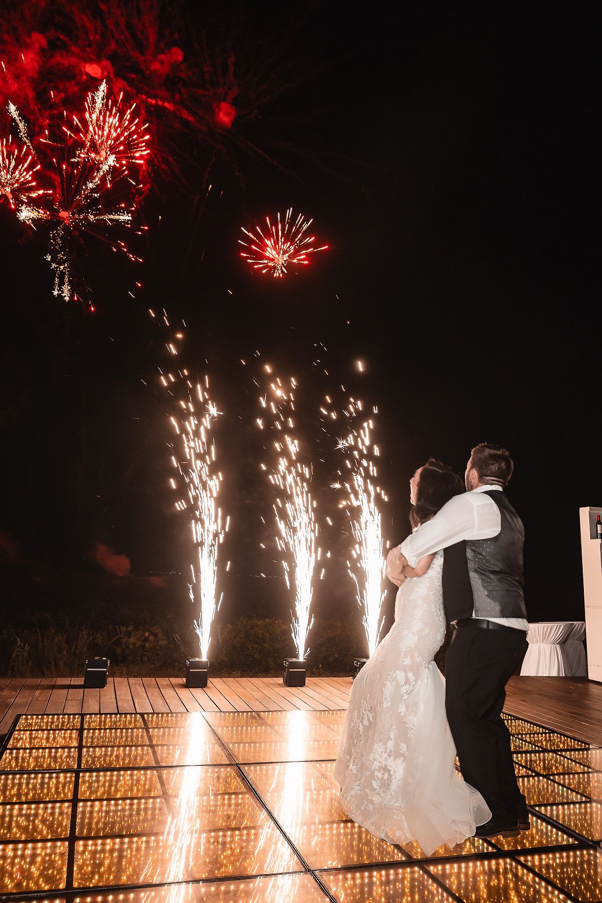 Bride and groom dancing on illuminated floor with stunning red and gold fireworks display at night wedding celebration.