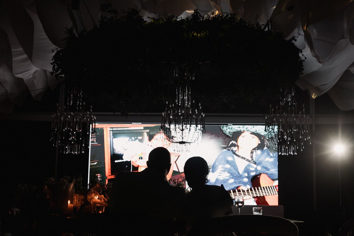 Silhouette of couple watching wedding highlight video on large screen during dimly lit reception.