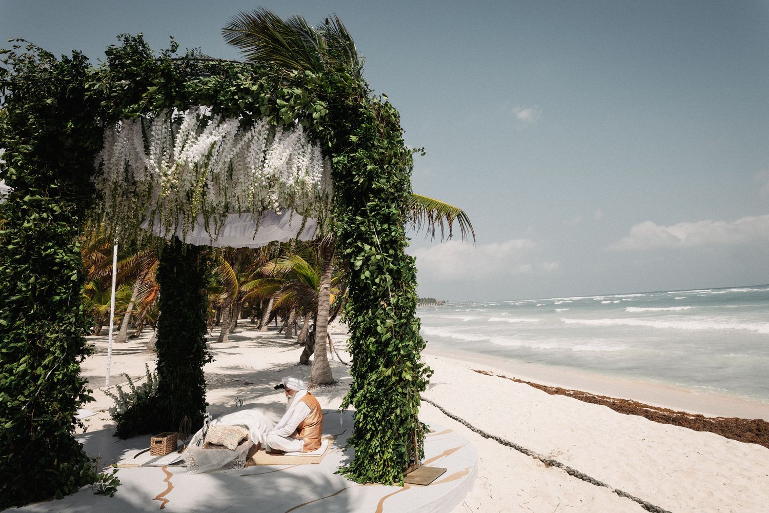 Couple relaxes under a lush green floral arch on a white sand beach with turquoise waves in Tulum, Mexico.
