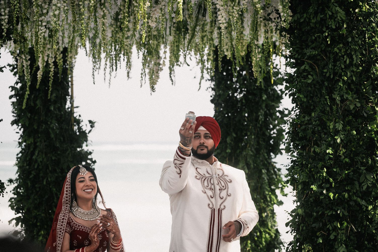 Sikh bride in red lehenga and groom in white sherwani celebrate joyfully under a white floral wedding arch.