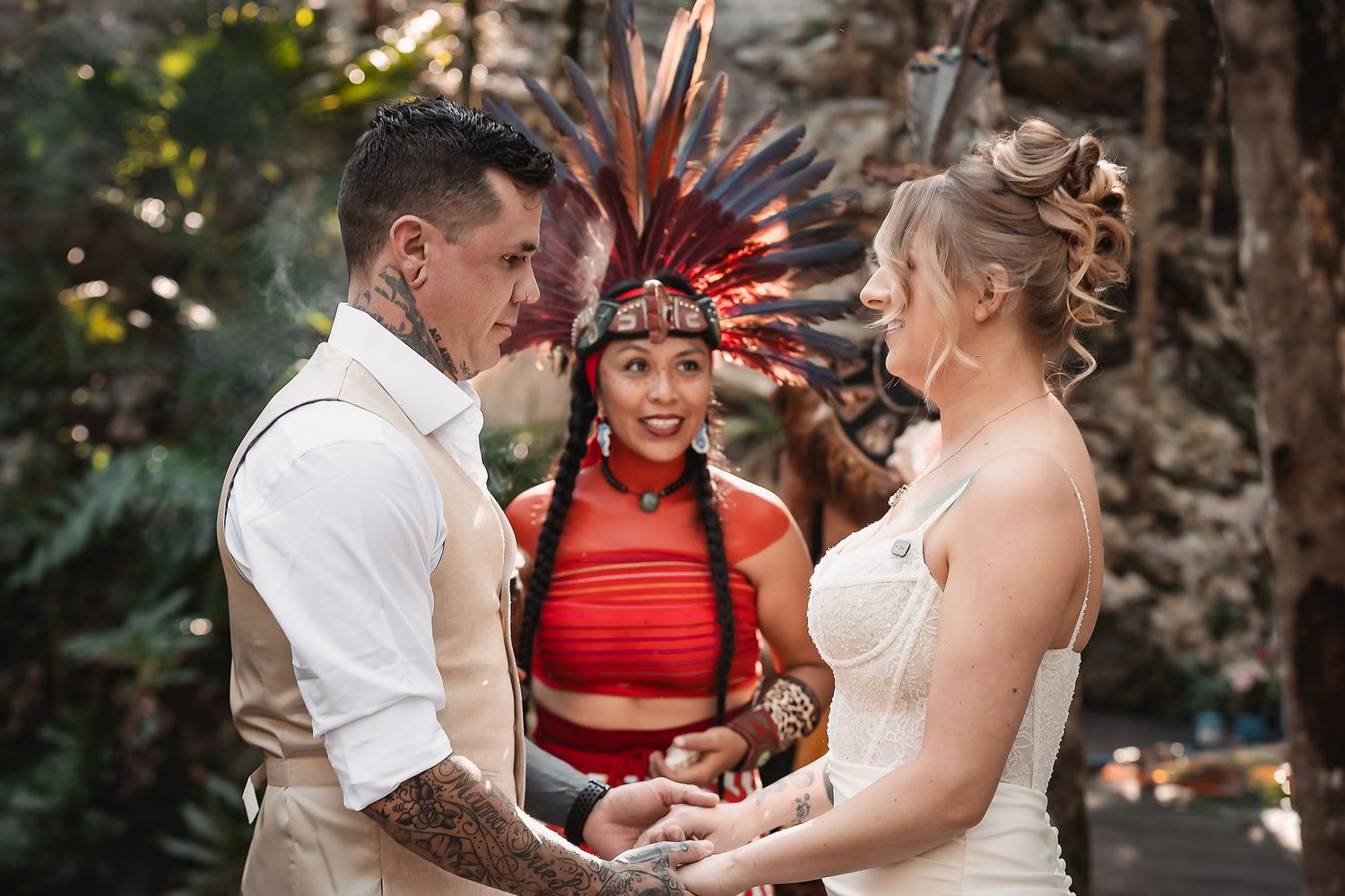 Couple holds hands during outdoor wedding ceremony officiated by woman in colorful Native headdress and red outfit.
