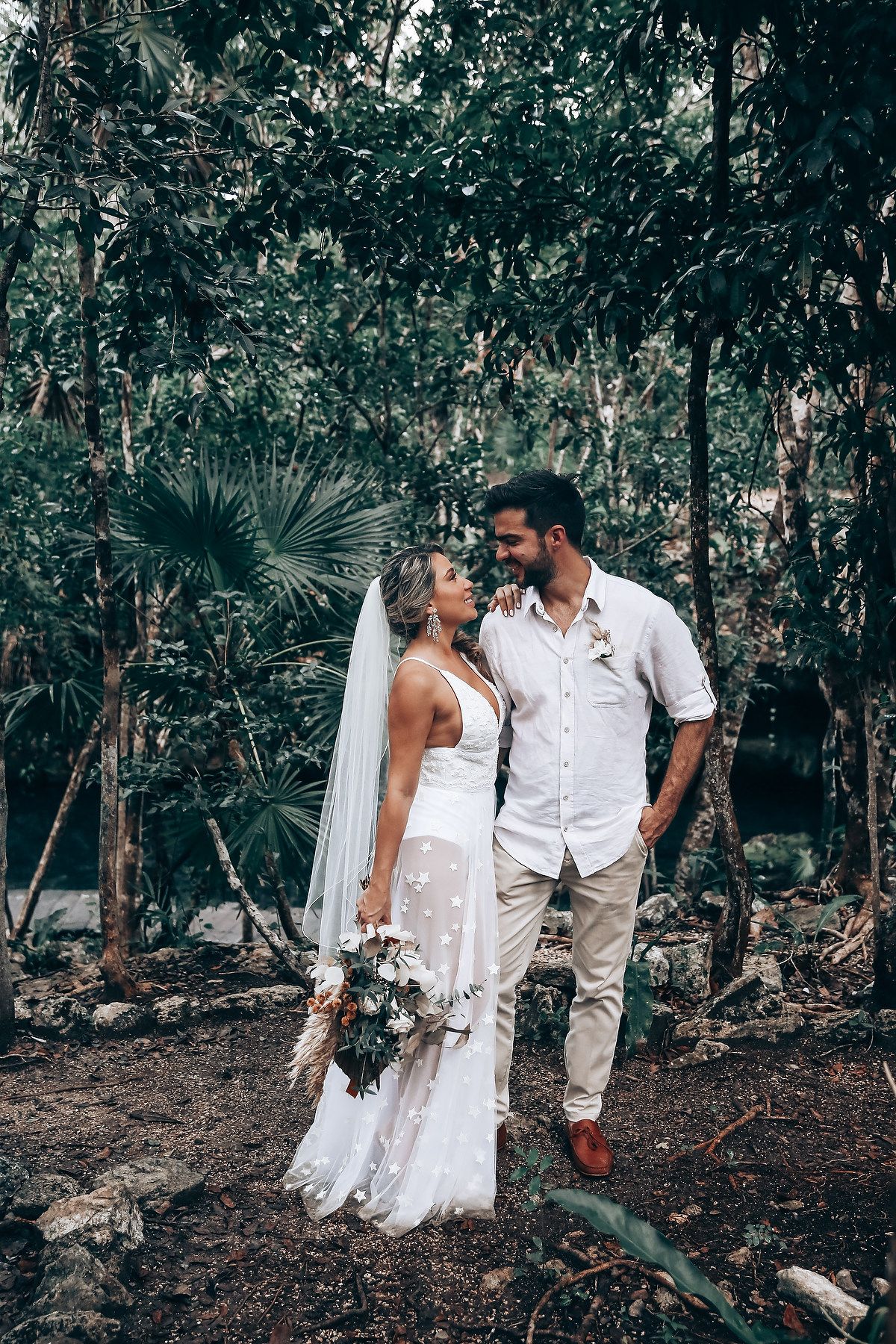 A couple in white attire poses together in a lush tropical forest setting.