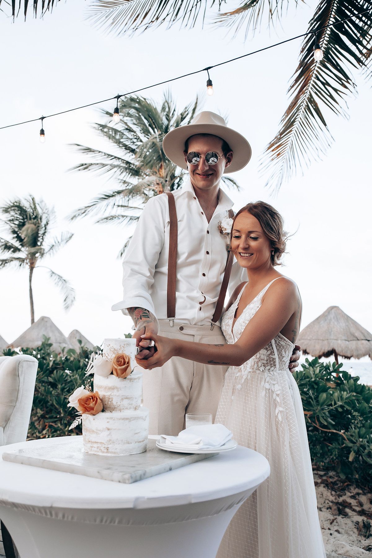 A couple cuts their wedding cake together at a tropical beach resort with palm trees in the background.