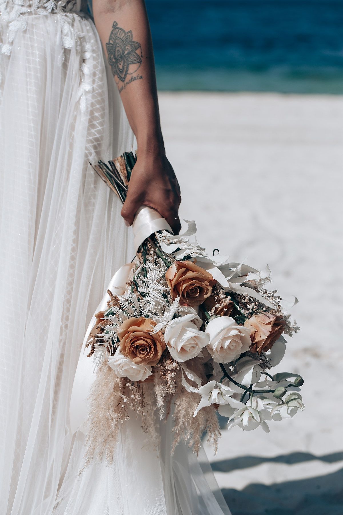 Elegant white and blush wedding bouquet with roses and dried flowers against a beachy blue background.