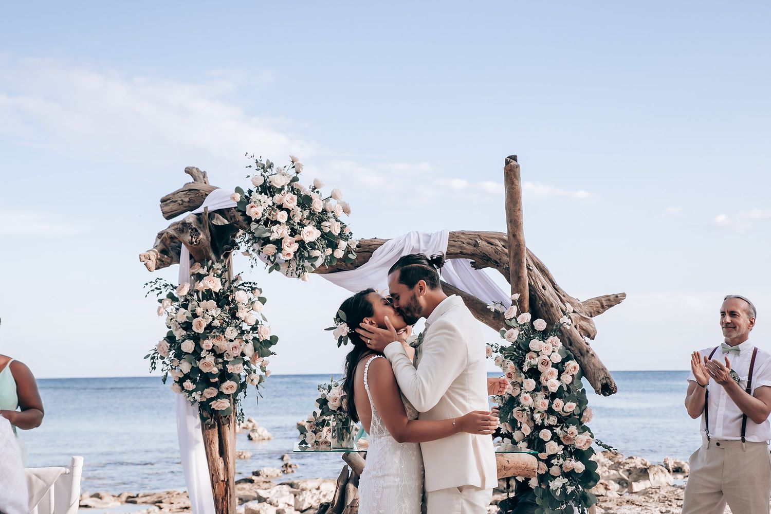 Wedding guests gather on a sunny beach with ocean views and tropical palm trees in the background.