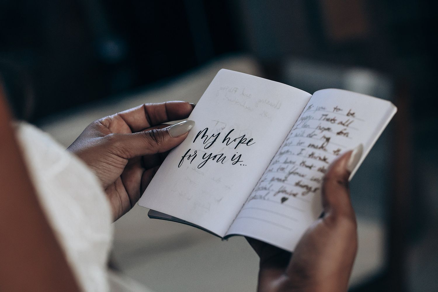 A person writing in a notebook with a pen on a dark surface, focusing on the handwritten text and paper.