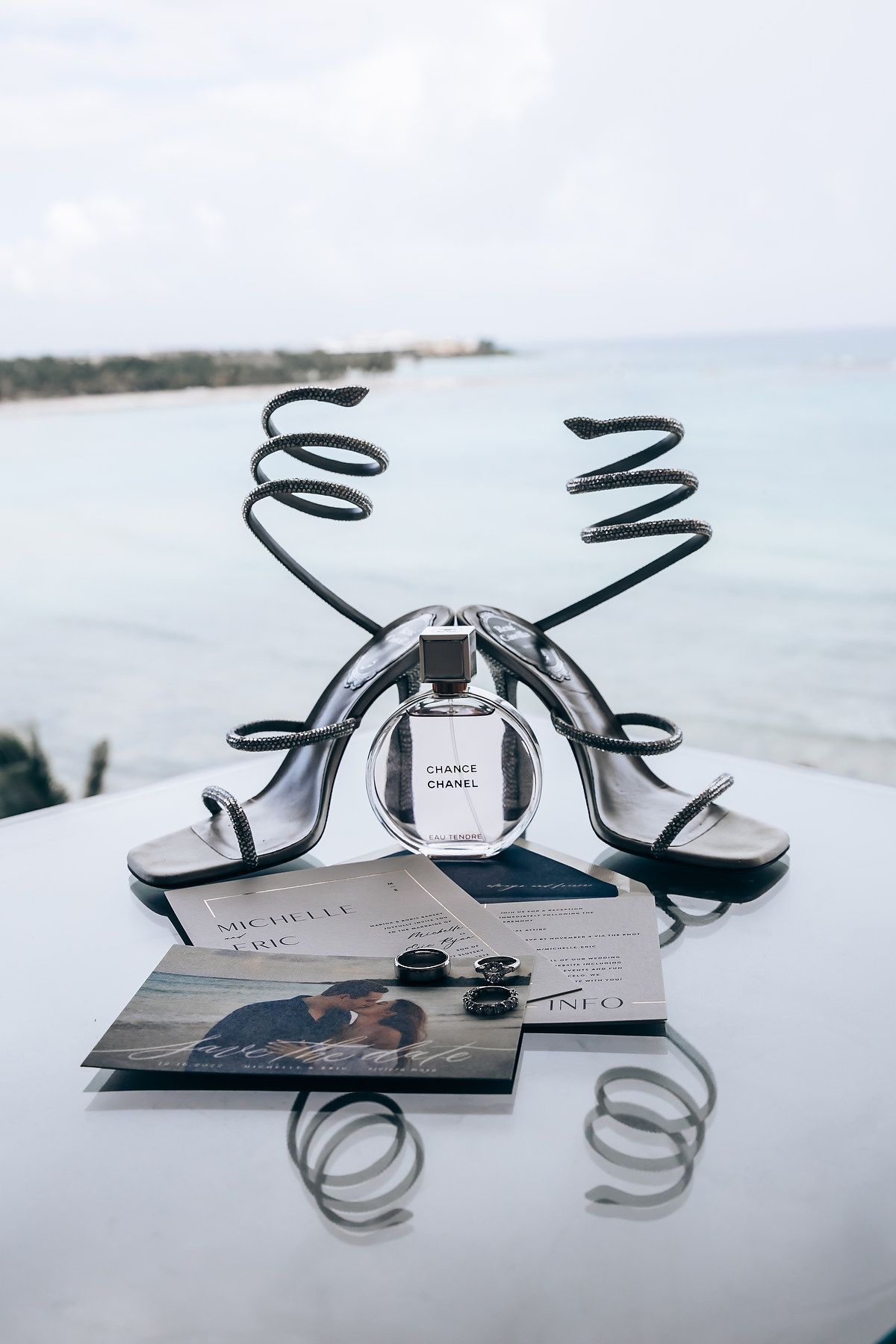 Wedding details flatlay with perfume bottle, sandals and photo cards against an ocean backdrop.