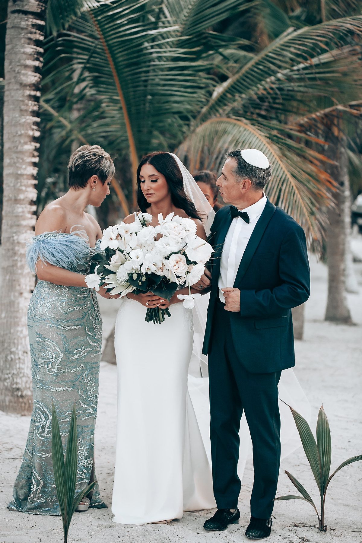 Elegant beach wedding scene with white bouquet and formal attire under palm trees.