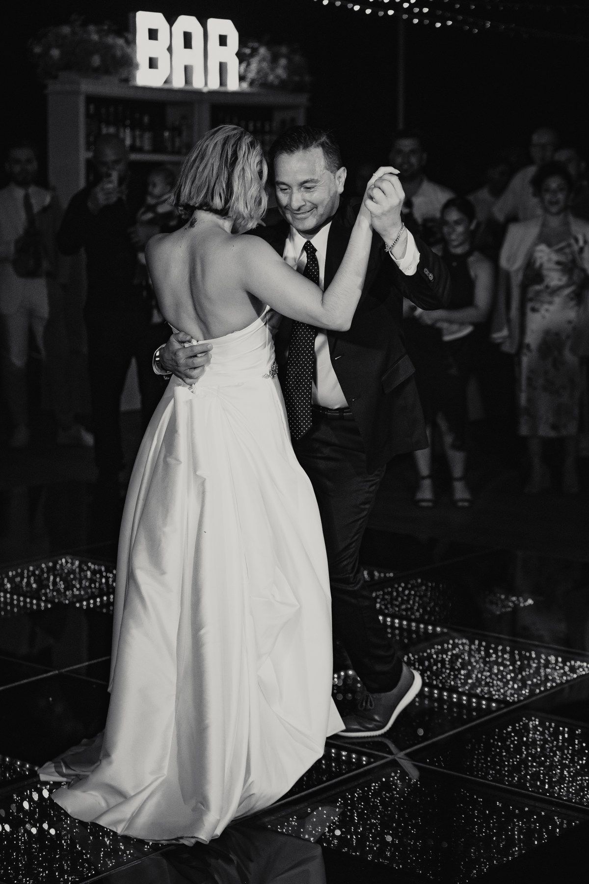 Black and white photo of bride and groom sharing romantic first dance at wedding reception near bar.