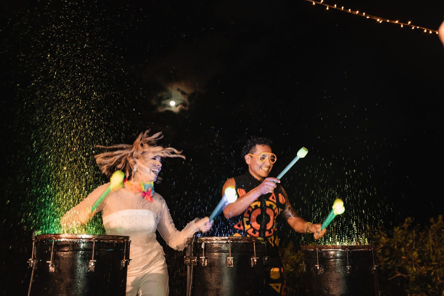 Two drummers perform energetically with glowing drumsticks at nighttime wedding reception.