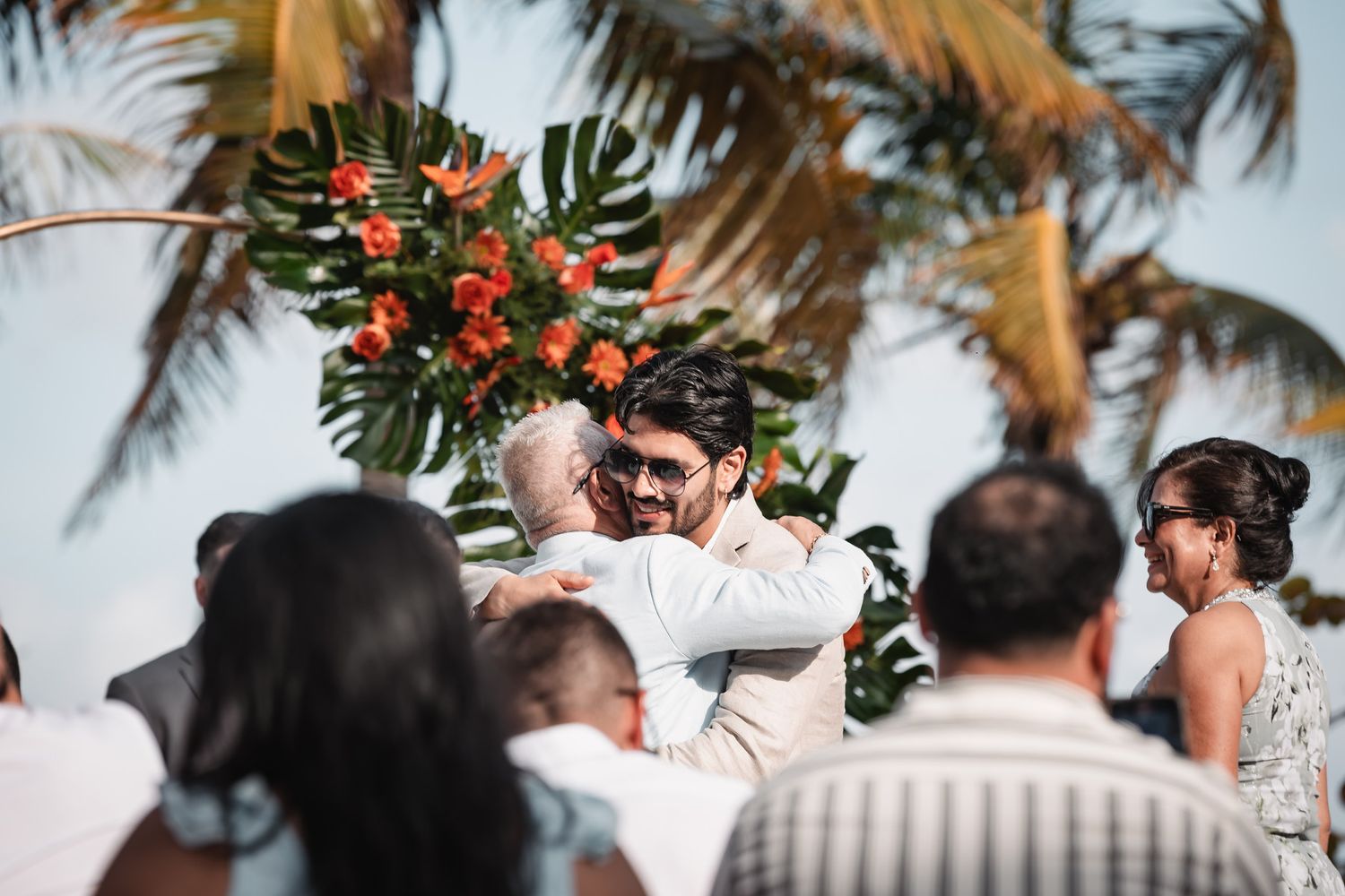 Groom embracing guest during tropical beach wedding ceremony with orange floral arch backdrop.