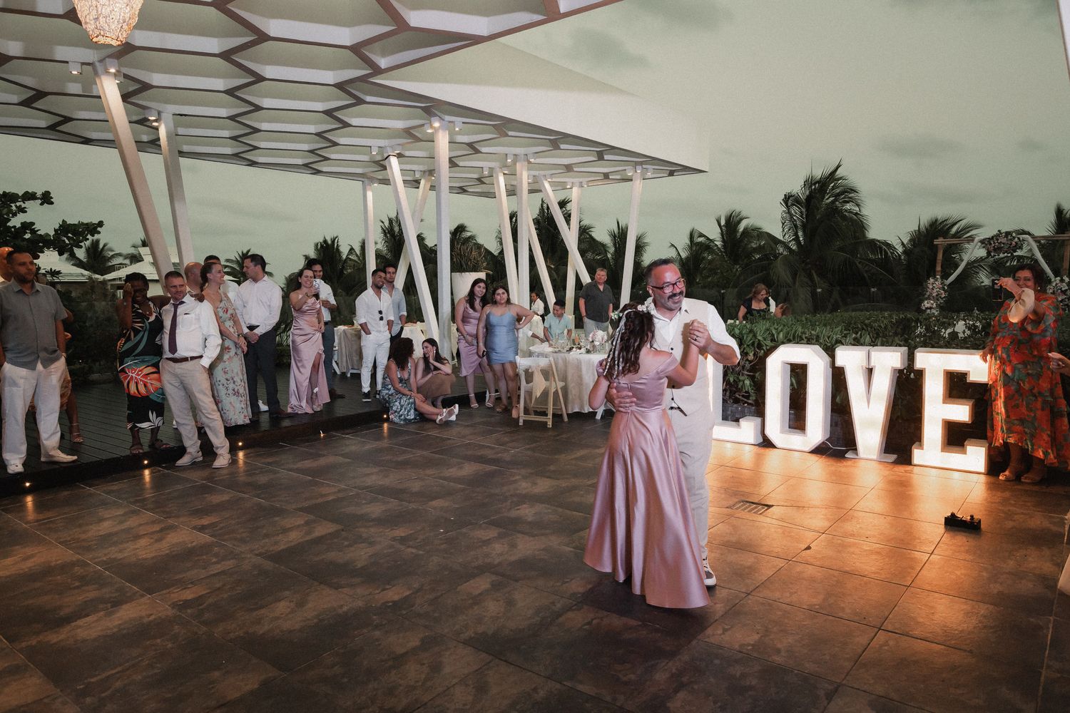 Couple shares first dance on outdoor terrace with illuminated LOVE letters and wedding guests at tropical venue.