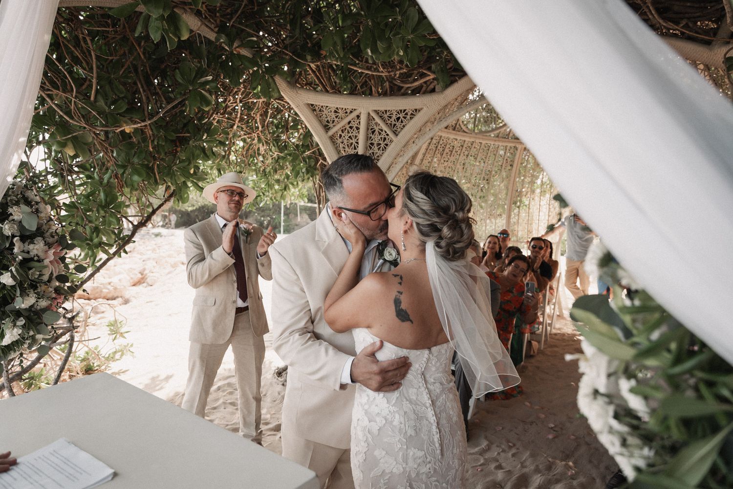 Newlywed couple shares first kiss under decorated arch at romantic outdoor beach wedding ceremony.