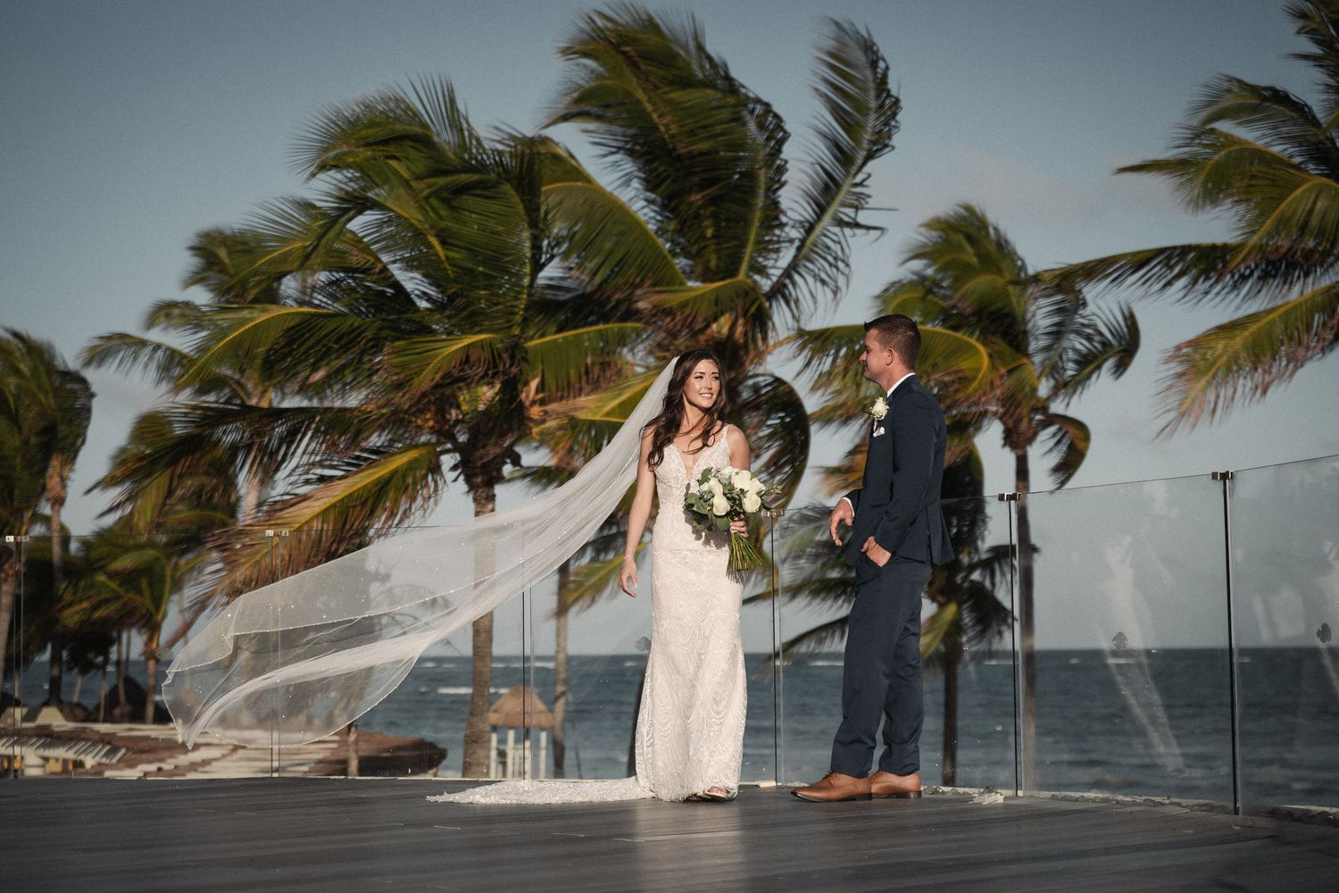 Bride and groom stand on oceanfront deck with dramatic wind-blown veil and swaying palm trees at sunset.