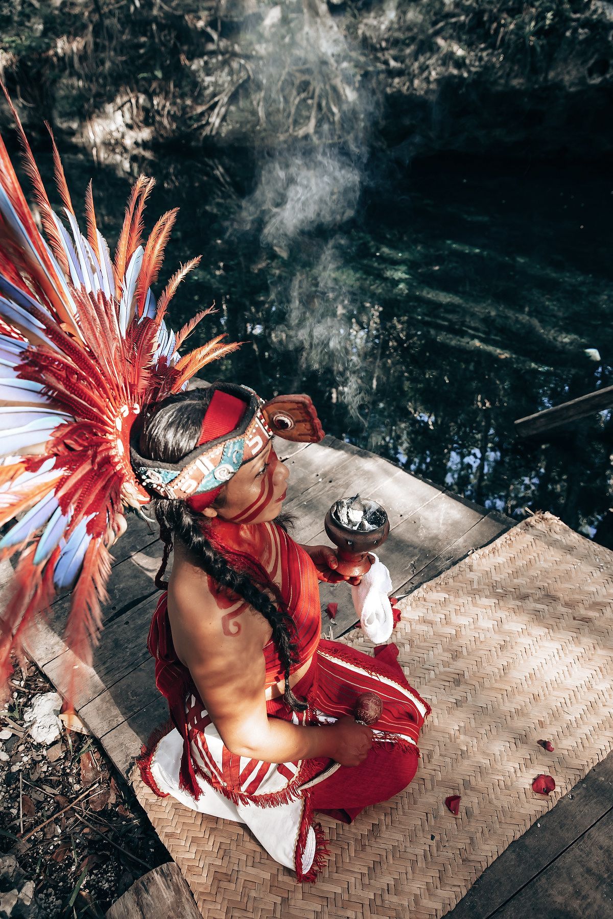 A person wearing a colorful feathered headdress and traditional clothing standing in a forest with trees and foliage.