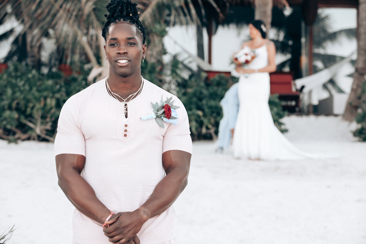 A smiling man stands in a tropical setting, with a woman in a wedding dress in the background.