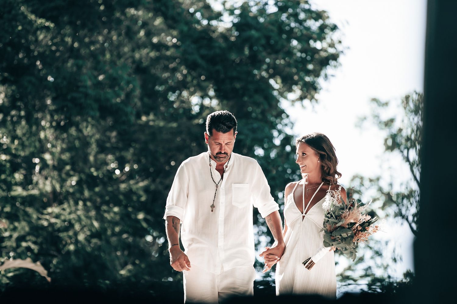 A couple in white clothing walking together in a park with trees in the background.