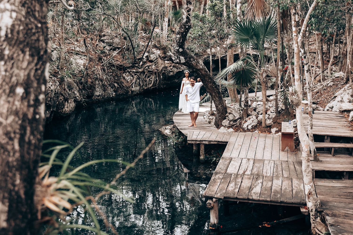 A wooden boardwalk winds through a tranquil cenote with dark blue waters surrounded by lush vegetation.