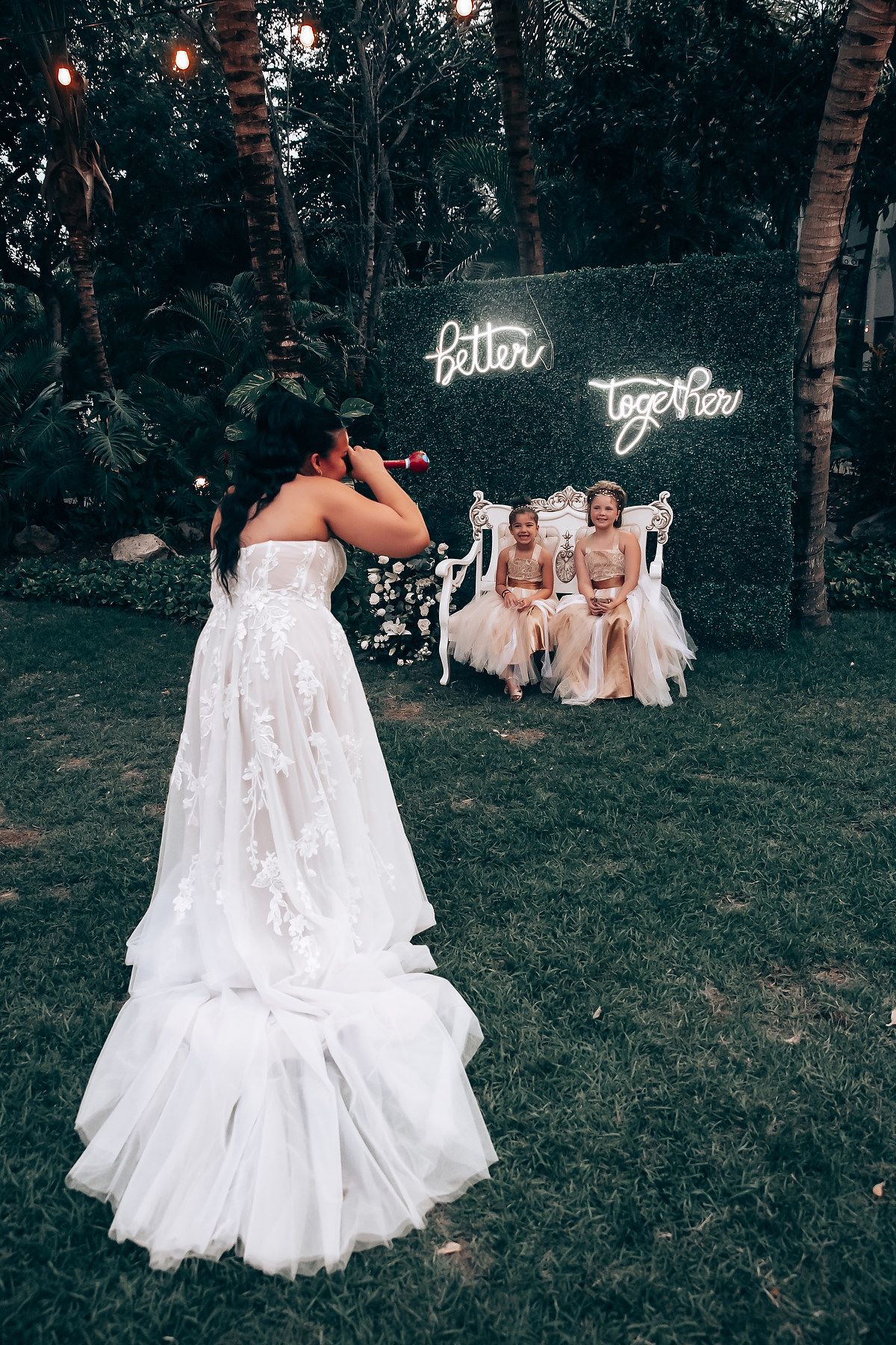 Wedding guests gather on a grassy lawn at dusk with glowing lights and white decorations creating a romantic atmosphere.