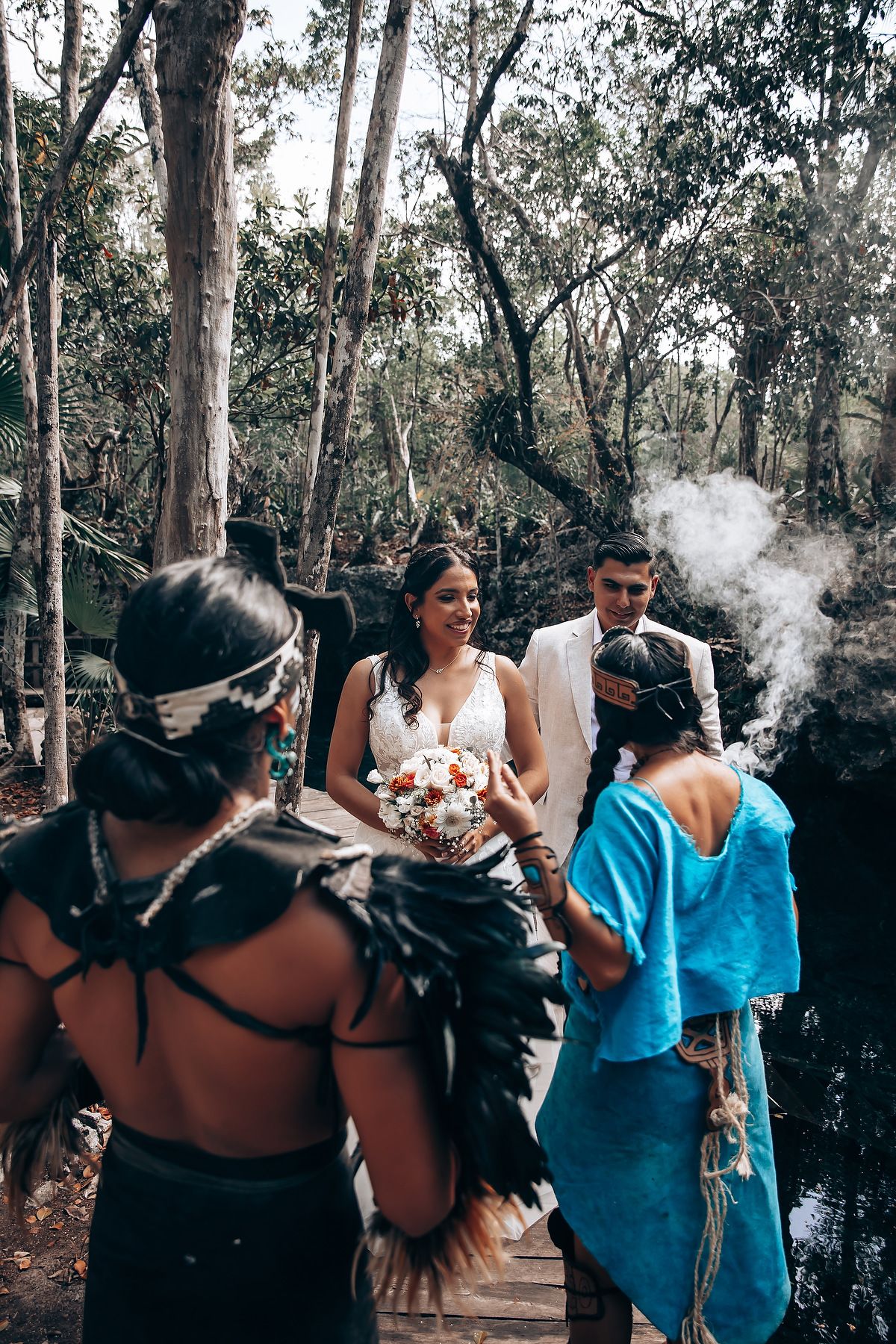 Wedding ceremony participants gather in a lush forest setting with smoke effects creating an atmospheric ambiance.