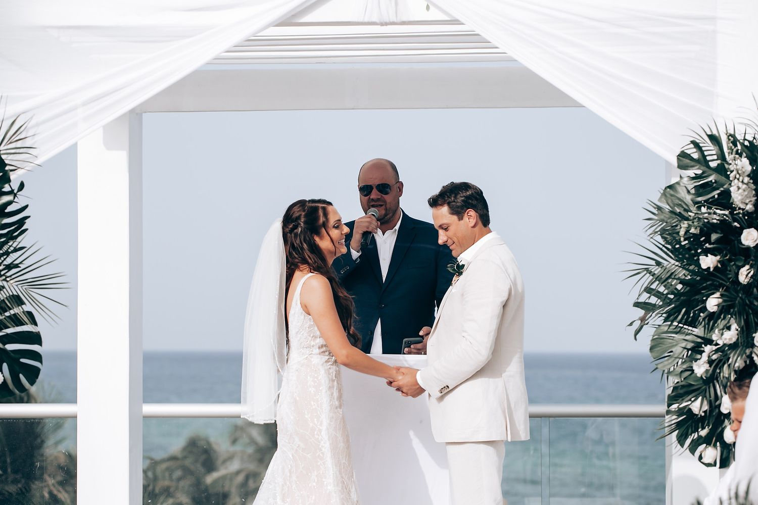 Intimate wedding ceremony under a white draped gazebo with ocean views and tropical decor.