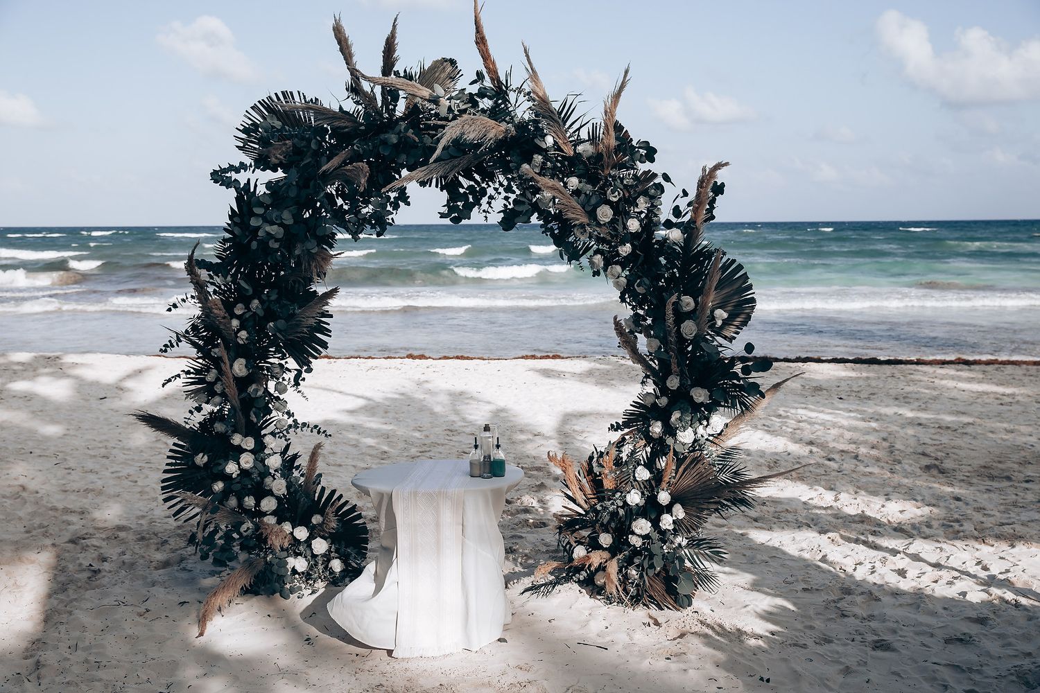 Beach wedding ceremony setup with floral arch and white decor against ocean waves at sunset.