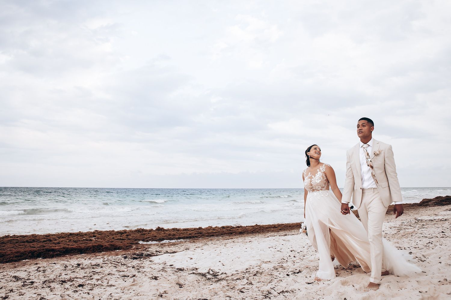 Couple walking along a serene beach with cloudy skies and ocean waves in the background.