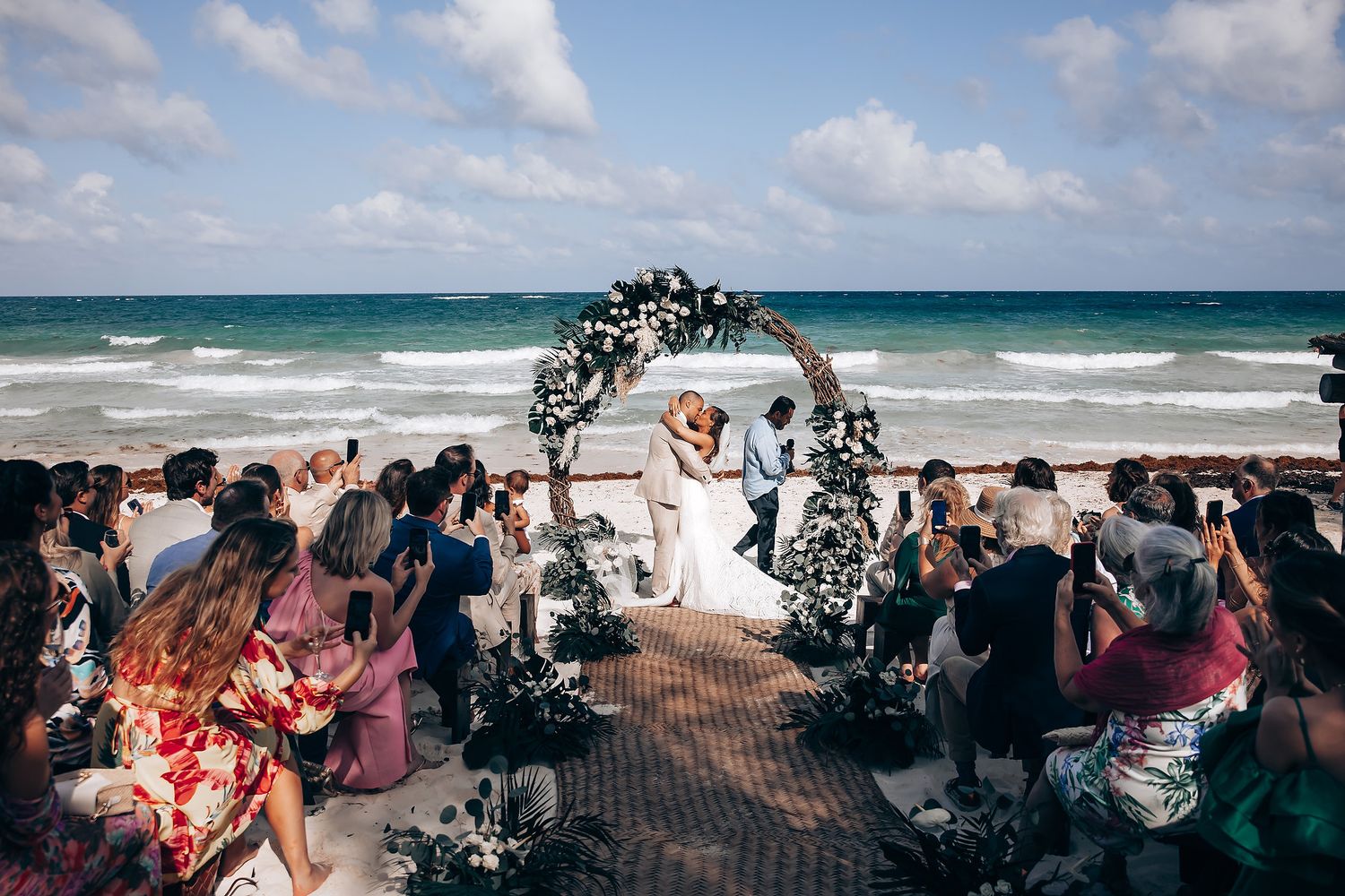 Wedding guests gather on a scenic beach with crashing waves and blue skies during an outdoor ceremony.