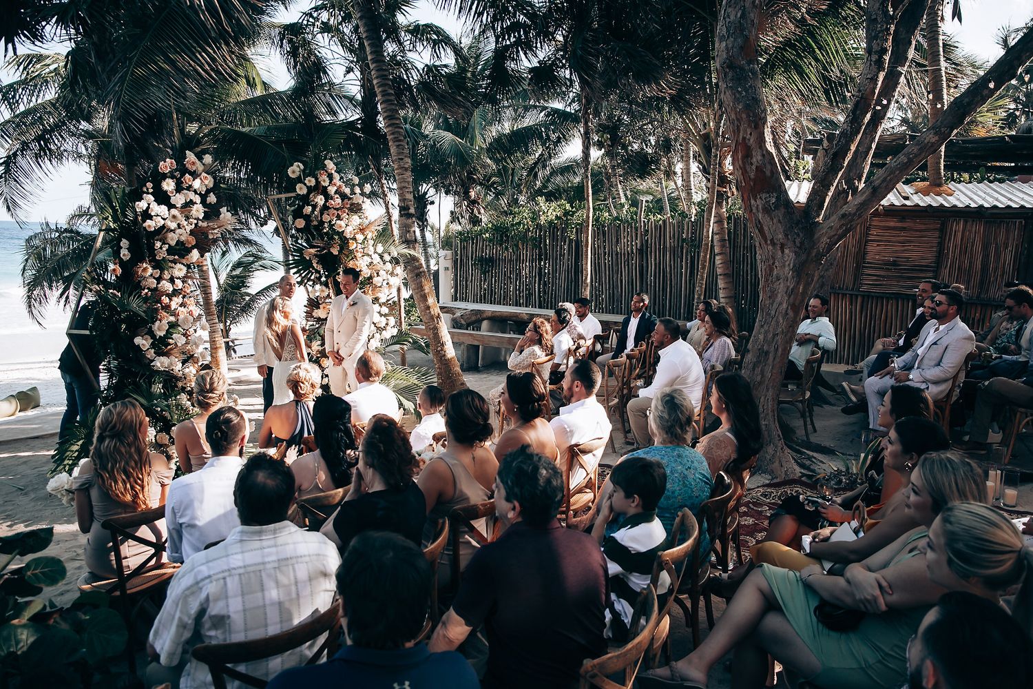 Large crowd of wedding guests seated outdoors under tropical palm trees during ceremony.