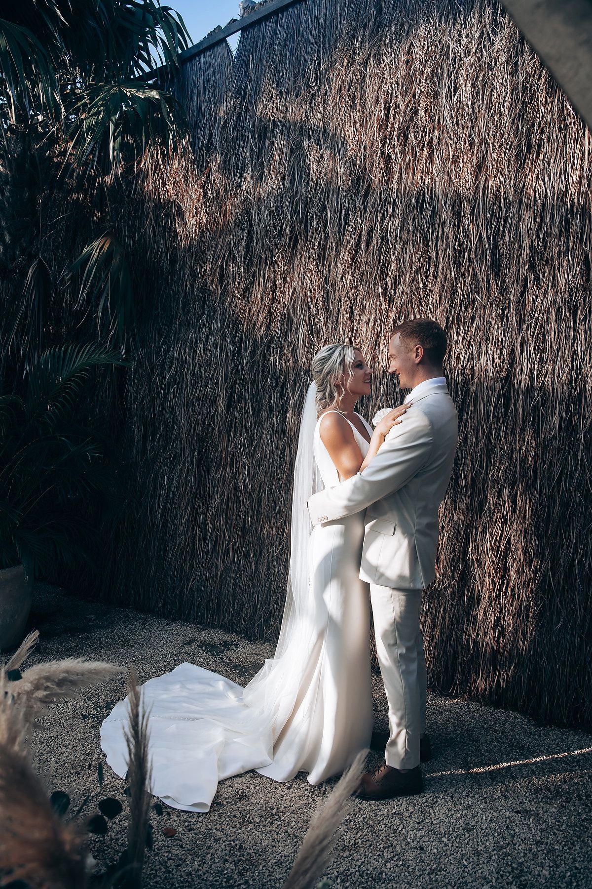 A romantic wedding moment against a rustic thatched wall backdrop with tropical palm leaves.