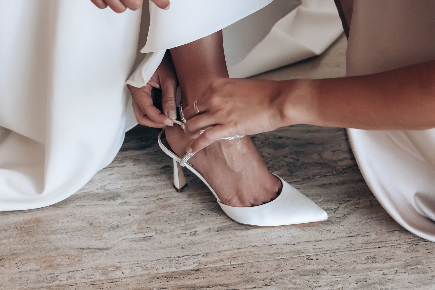 Close-up of hands fastening a white bridal shoe strap on wooden flooring.