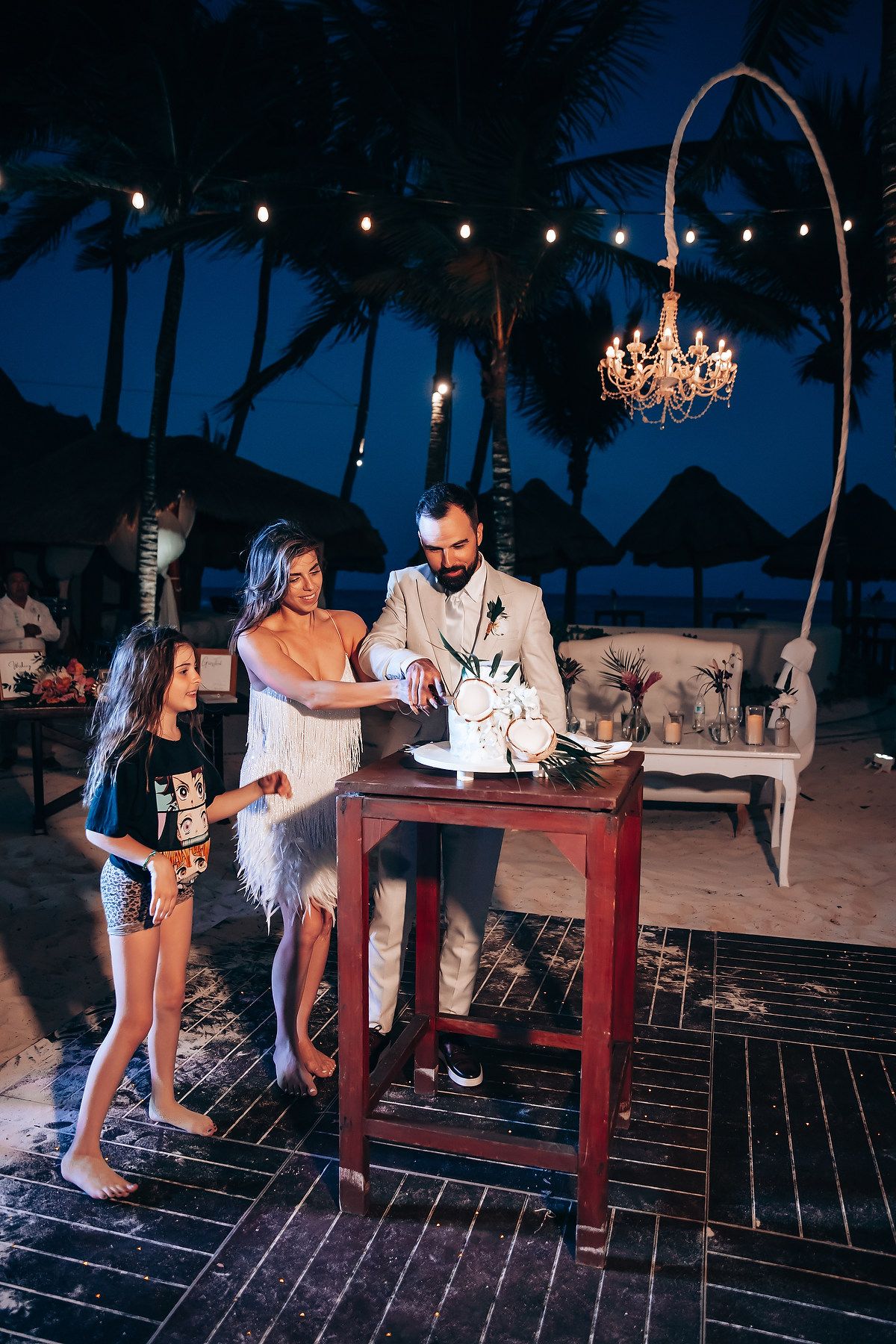 Guests enjoying drinks at a wooden bar setup during a nighttime beach wedding reception.