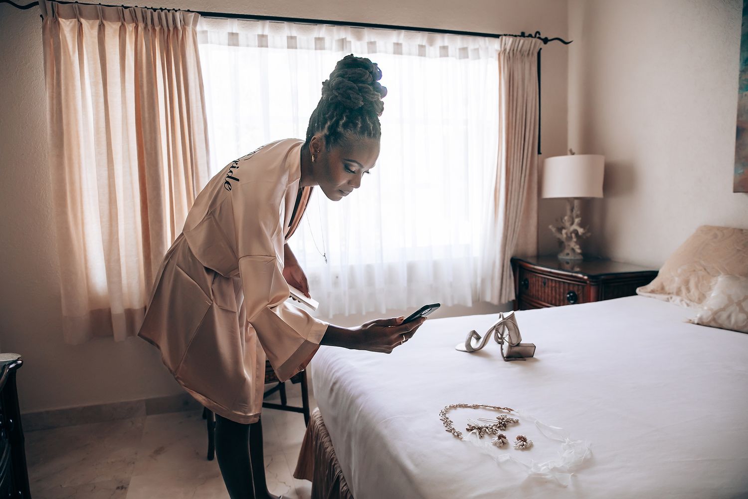 Wedding preparation scene showing someone making final touches to a white bed in a sunlit hotel room.