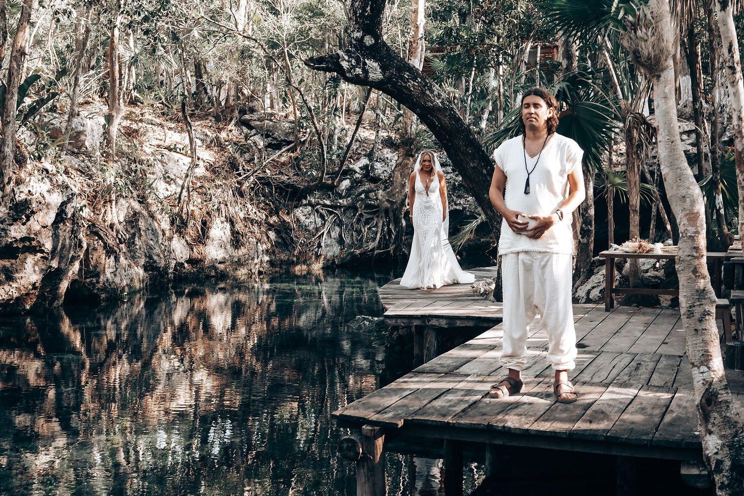 A couple in white outfits walks through a wooded area with Spanish moss hanging from trees during a photo session.