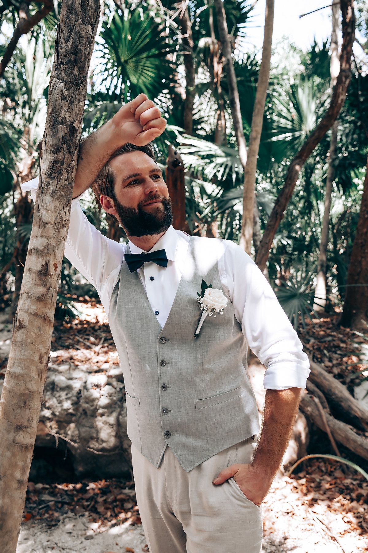 Groom in light gray vest and bowtie poses casually among tropical palm trees.