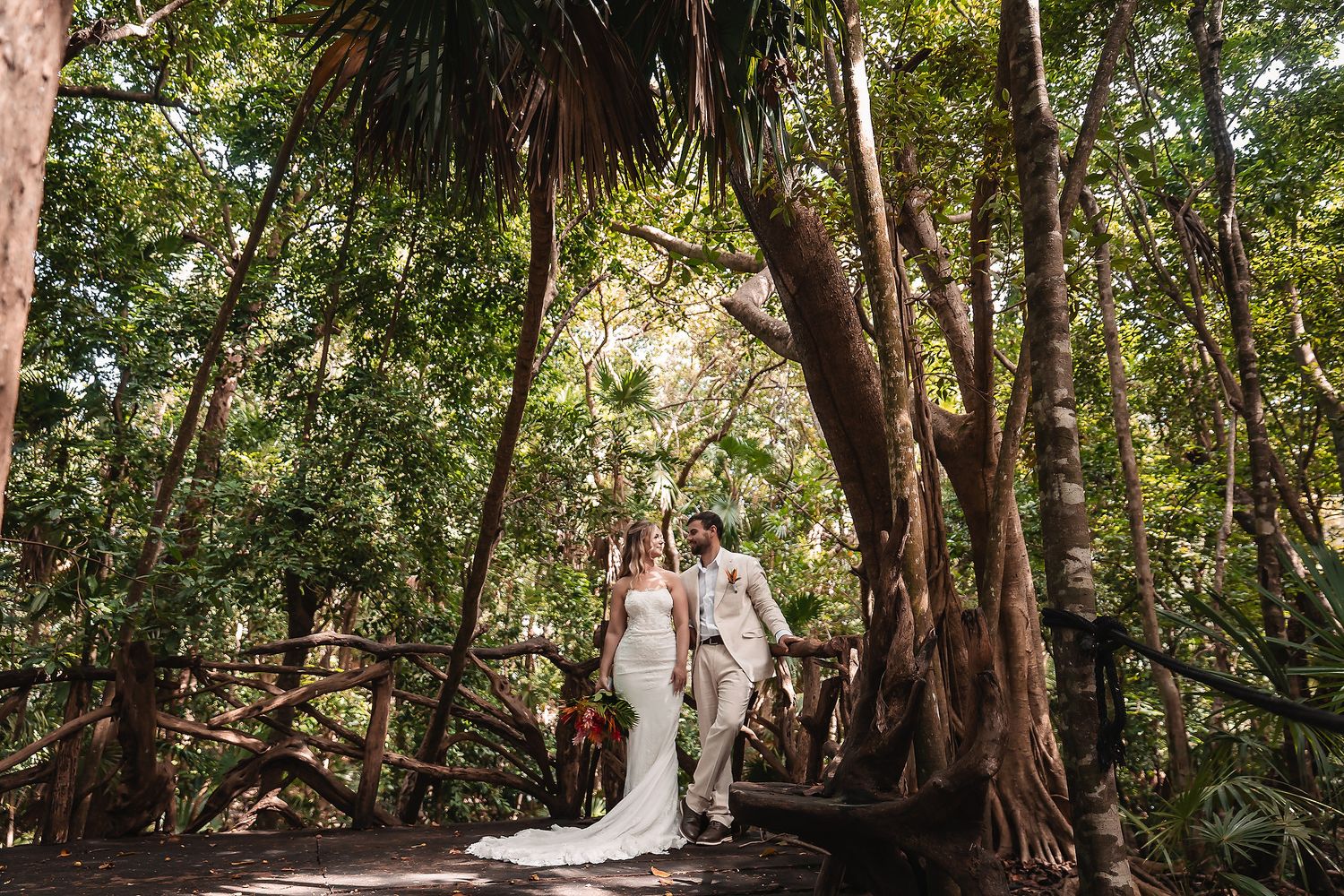 Bride and groom walk hand-in-hand on a wooden bridge surrounded by lush tropical forest trees.
