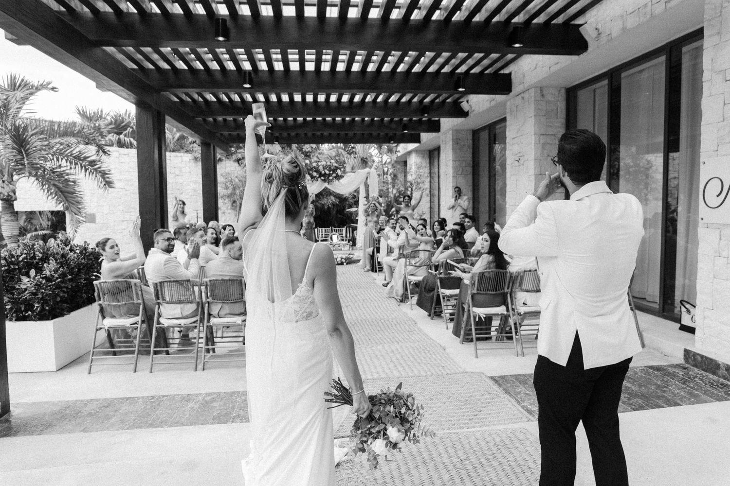 Black and white photo of bride and groom entering outdoor wedding ceremony with guests seated under pergola.