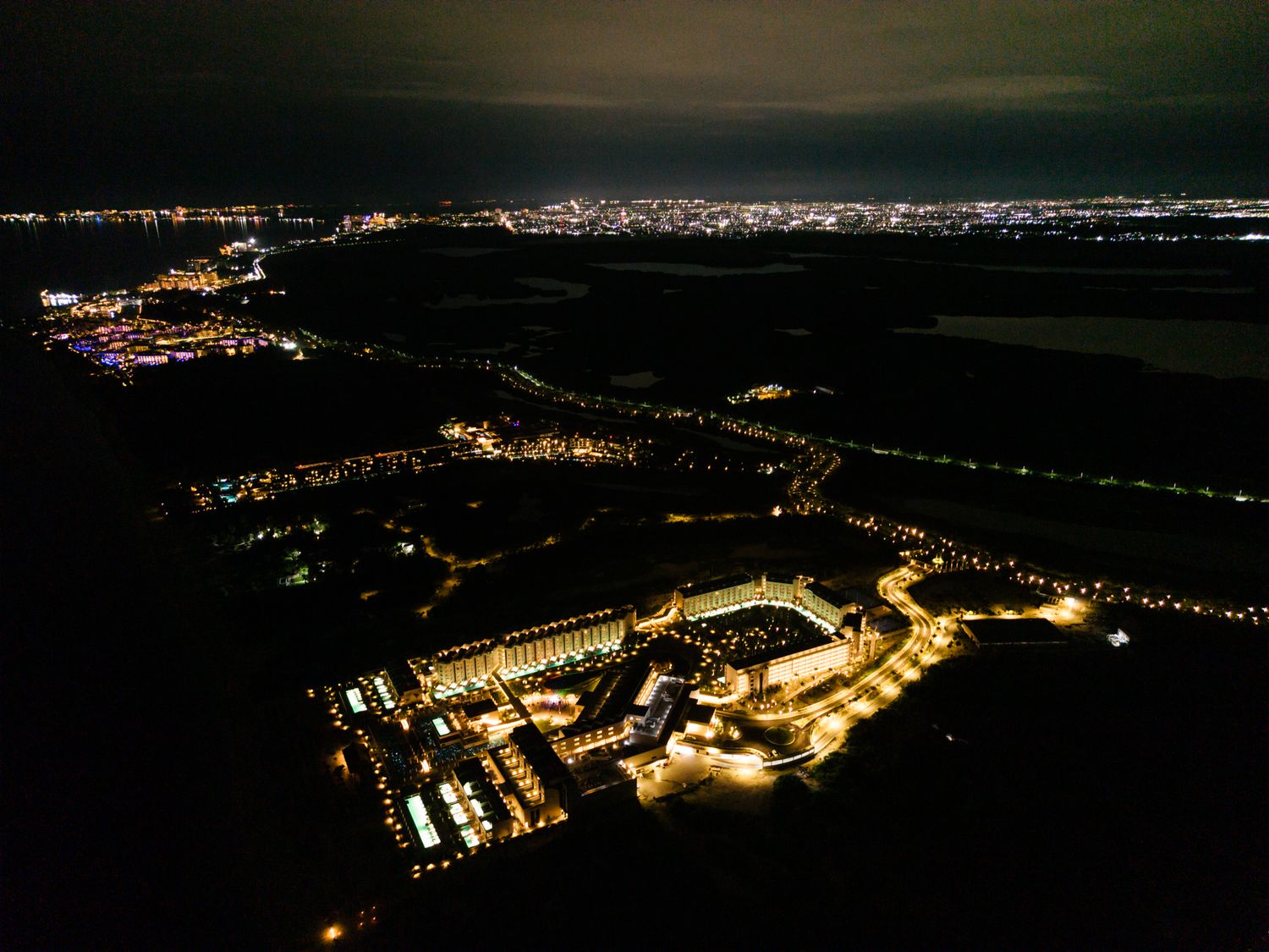 Aerial night view of a illuminated coastal town with glowing city lights reflecting on dark water in the distance.