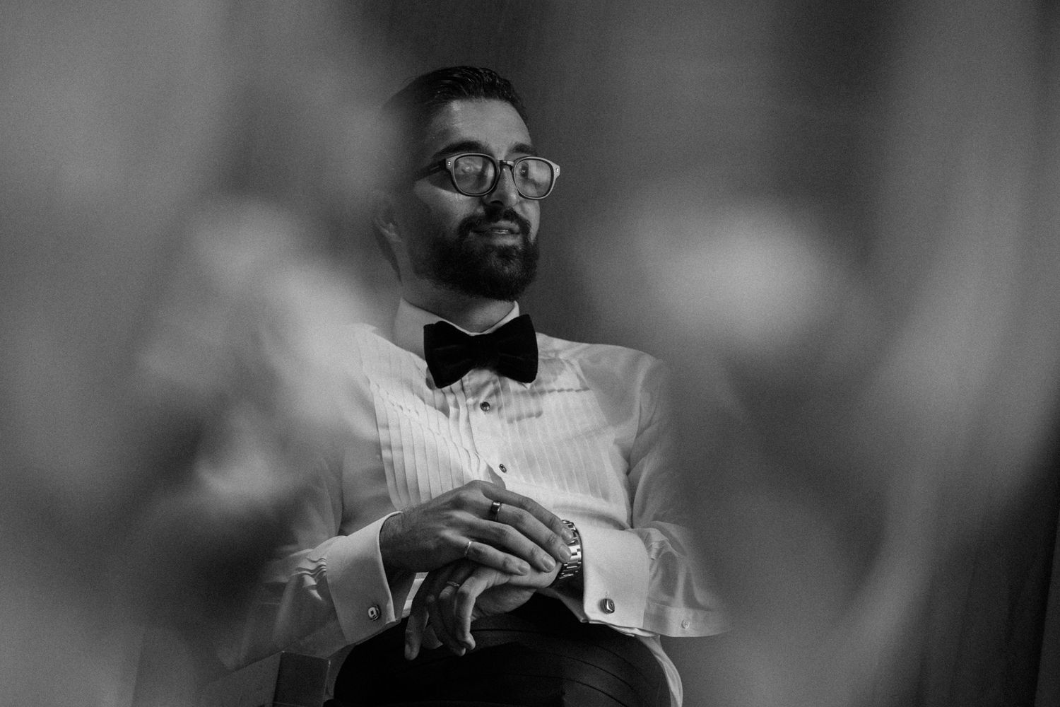 Black and white portrait of a bearded man in a tuxedo with bow tie and glasses, sitting thoughtfully.