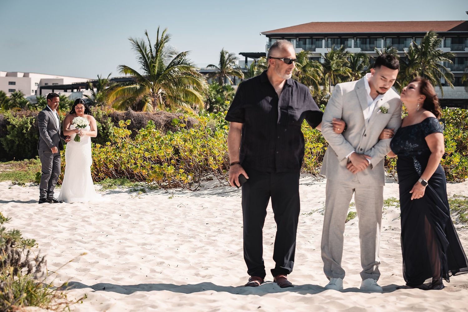 Groom embracing family on sandy beach wedding venue with bride and palm trees in background.