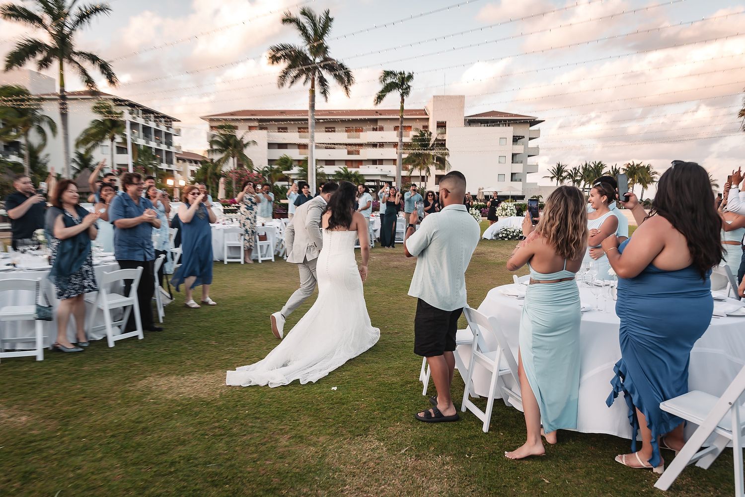 Bride walks through cheering wedding guests at an outdoor beachside reception with palm trees.